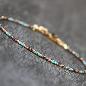 Op de afbeelding: Delicate armband met kleine, vierkante kralen in goud, bruin en turquoise. De armband heeft een goudkleurige sluiting. Een minimalistisch sieraad, geschikt voor dagelijks gebruik. Lengte ongeveer 18 cm.
