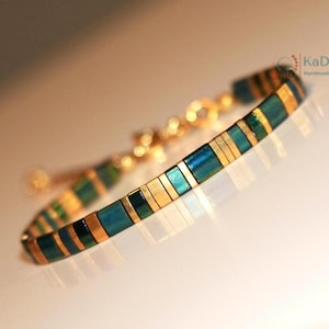 May include: A gold-toned bracelet with rectangular teal and gold segments. The bracelet has a delicate chain and clasp. The handmade jewelry piece is displayed on a white surface.