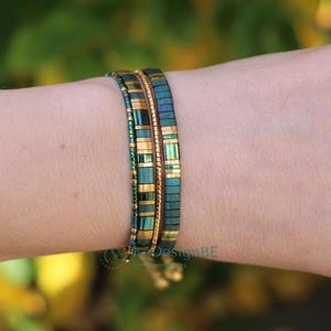 May include: Two beaded bracelets are worn on a wrist. The bracelets feature a teal and gold color scheme. The beads are rectangular and arranged in a linear pattern. The bracelets are a stylish accessory.
