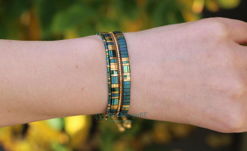 May include: Two beaded bracelets are worn on a wrist. The bracelets feature a teal and gold color scheme. The beads are rectangular and arranged in a linear pattern. The bracelets are a stylish accessory.