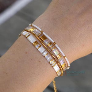 Peut inclure: Plusieurs bracelets de perles blanches et dorées superposés sur un poignet. Les bracelets présentent des perles rectangulaires et rondes. Les accents dorés ajoutent une touche d'élégance. Un accessoire élégant.