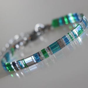 May include: A close-up of a bracelet with a silver chain and a central band of rectangular beads. The beads alternate between silver, teal, blue, and green. The bracelet is on a reflective surface.