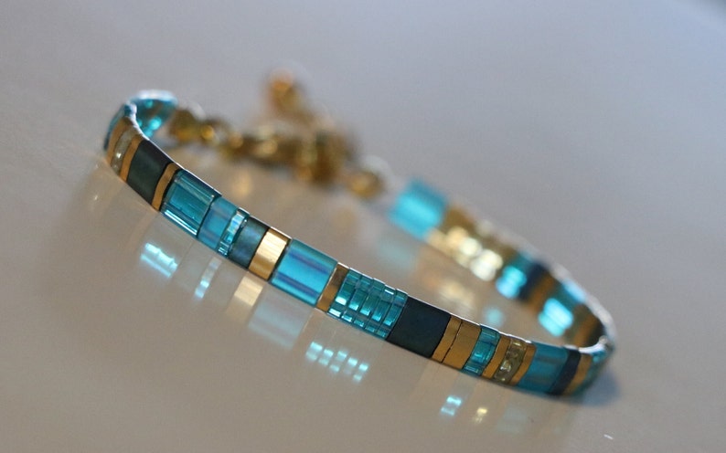 tila bracelet close up, flats square beads in aqua, gold, blueish beads in random pattern.