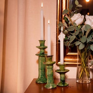 Puede incluir: Tres candelabros de cerámica verde con velas blancas encendidas. Los candelabros son de diferentes alturas y tienen un aspecto texturizado y rústico. Un jarrón con rosas blancas y vegetación es visible en el fondo.