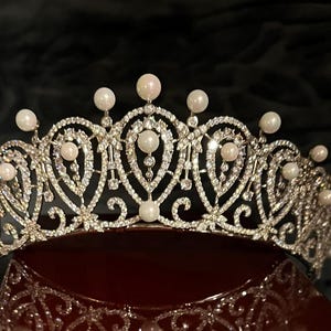 May include: A silver-toned tiara with a delicate design, adorned with numerous clear crystals and creamy-white pearls. The tiara is displayed on a red surface against a dark background, showcasing its intricate details and elegant style.