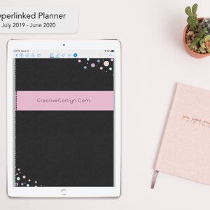Può includere: Un planner digitale nero e rosa su una superficie bianca con una piccola pianta grassa in vaso e un quaderno rosa con il testo "you were made for this" in lettere dorate. Il planner è aperto su una pagina con il testo "CreativeCaitlyn.Com" e un titolo che dice "Hyperlinked Planner July 2019 - June 2020".