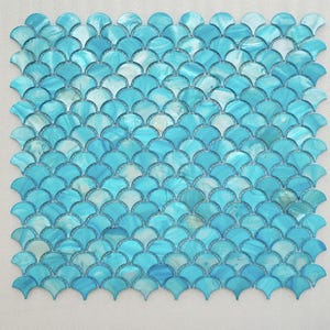 May include: A square mosaic tile sheet with a pattern of overlapping, fan-shaped tiles. The tiles are a mix of light and medium blue, with some white accents, creating a watery effect. The design resembles mermaid scales.