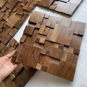 May include: Wooden wall panels with a 3D geometric design. The panels are made of dark brown wood and feature a variety of square and rectangular blocks arranged in a textured pattern. The panels are shown on a white surface. The panels are made of dark brown wood.