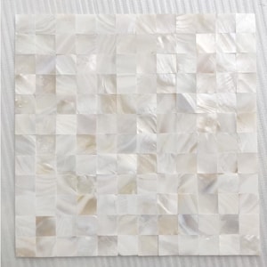 Könnte beinhalten: Quadratische Mosaikfliesenplatte mit Perlmutt-Finish. Die Fliesen sind in einem Rastermuster angeordnet und erzeugen eine schimmernde, irisierende Oberfläche. Die Farbpalette ist überwiegend weiß mit subtilen Variationen von Beige und Creme.