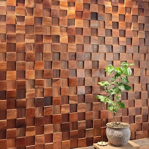 Puede incluir: Una pared de madera compuesta por numerosos bloques cuadrados pequeños en tonos marrones variados. Los bloques crean un efecto texturizado tridimensional. Una pequeña planta en maceta se encuentra frente a la pared, sobre una superficie de madera.