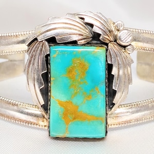 May include: A silver cuff bracelet featuring a rectangular turquoise stone with golden-brown matrix patterns. The center stone is flanked by decorative silver leaf motifs and small silver spheres, set on a split-shank band with beaded edges.