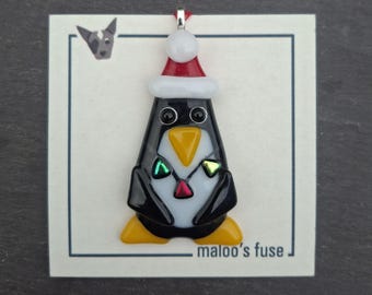 Könnte beinhalten: Ein Anhänger aus geschmolzenem Glas in Form eines Pinguins mit schwarz-weißem Körper, gelben Füßen und Schnabel sowie einer rot-weißen Weihnachtsmütze. Der Pinguin trägt eine Kette mit drei kleinen dreieckigen Anhängern auf einer Karte mit maloo's fuse.