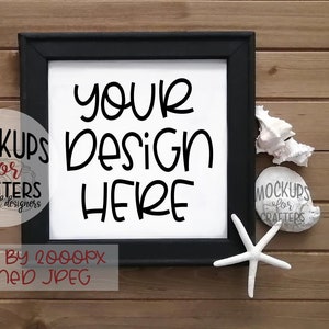 Op de afbeelding: Een zwarte geframed canvas mockup met de tekst "YOUR DESIGN HERE" op een witte achtergrond. De canvas ligt op een houten oppervlak met schelpen en een stervormige zeester.