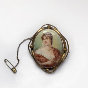 May include: A gold-toned oval brooch featuring a painted portrait of a woman in a red dress. The brooch has a safety pin attached to a chain.