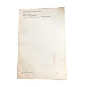 May include: A worn book cover titled "DADA AND SURREALISM REVIEWED" by Dawn Ades. The cover is off-white with black text and a pink streak. Published in 1978 by the Arts Council of Great Britain, it outlines Dada and Surrealism.