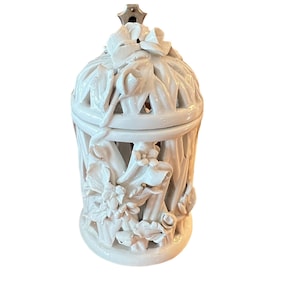 May include: A white ceramic lantern with a floral design. The lantern has a cage-like structure with cut-out patterns and is adorned with sculpted flowers and leaves. A metal fixture is attached to the top.