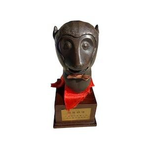 May include: A bronze monkey head sculpture with a bow tie and red fabric. The sculpture is mounted on a wooden base with a gold plaque. The monkey has detailed facial features and is a unique decorative item.