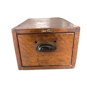 May include: A rectangular brown wooden storage box with a single drawer featuring a dark metal cup pull handle. A small rectangular metal label plate with the text "Globe-Wernicke" is attached to the upper front frame of the wooden unit.