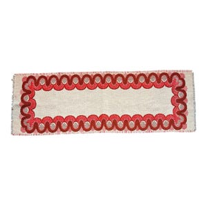 May include: Rectangular beige table runner with a scalloped red border. The border features a repeating pattern of interlocking circles in varying shades of red. The edges are fringed.
