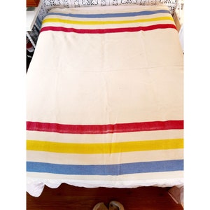 May include: Cream-colored blanket with horizontal stripes in red, yellow, and blue. The blanket is spread across a bed, showcasing its simple yet striking design. The stripes are evenly spaced, creating a balanced and visually appealing pattern.
