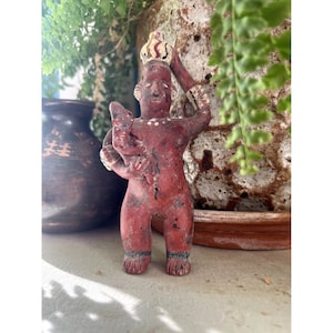 May include: A small, red-painted figurine of a standing figure holding a smaller figure. The figurine has a crown and armbands. The figure is standing in front of a dark brown vase and green plants.