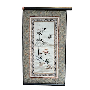 May include: An embroidered wall hanging featuring a bamboo stalk, flowers, and a bird. The artwork is framed by a decorative border with floral and geometric patterns. The color palette includes shades of green, red, and cream, with a dark wooden rod at the top.