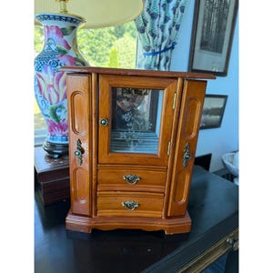May include: A wooden jewelry armoire with a warm brown finish, featuring a central glass door with an etched floral design, two lower drawers with ornate metal pulls, and two side panels with vertical recessed detailing and decorative hardware.