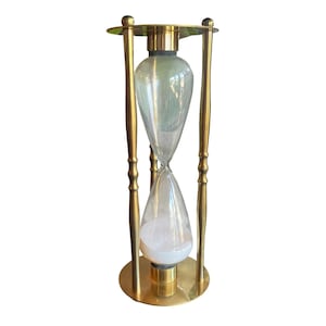 May include: A tall hourglass featuring a polished gold-tone metal frame with four slender vertical supports connecting a circular top and base. The central glass vessel contains white sand, with the majority of the sand settled in the bottom bulb.