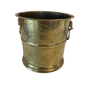 May include: A vintage brass bucket with etched Asian-inspired designs. The bucket features a cylindrical shape with a flared rim and two ring handles. The exterior is adorned with cloud and architectural motifs.