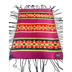 May include: A magenta-colored blanket with a geometric pattern in orange, yellow, green, and white. The blanket has black fringe along the edges. The design includes horizontal stripes and diamond shapes.