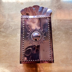 May include: A copper-colored metal wall pocket with a sunburst design and a scalloped top. The rectangular pocket is adorned with small, evenly spaced rivets along the edges. The sunburst has a central, raised circle surrounded by radiating lines.
