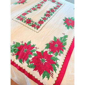 May include: Cream-colored rectangular tablecloth with a red and green poinsettia floral design. The border is red with a scalloped edge. The center features a rectangular floral pattern, and the corners have large poinsettia arrangements.