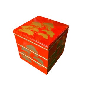 May include: A stackable, red lacquered Japanese bento box with gold-colored floral designs. The box has a square shape and is divided into three sections. The gold designs are on the top and sides of the box.