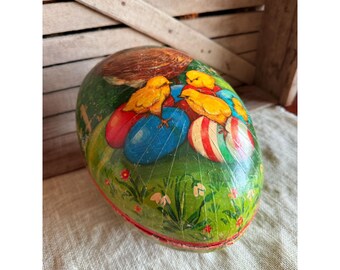 May include: An egg-shaped container with a painted Easter theme. The container features yellow chicks, colorful eggs, and a green grassy background. The egg is decorated with red, blue, and white stripes.