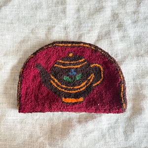 May include: A handmade, half-circle tea cozy featuring a brown teapot design on a burgundy background. The teapot has orange and brown accents, with a small blue and green floral detail. The cozy is made of textured yarn and has a brown border.