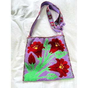 May include: A purple shoulder bag with a floral design. The bag features large red flowers with yellow centers and green stems and leaves. The strap is purple with a red and green pattern. The bag is made of a woven fabric.