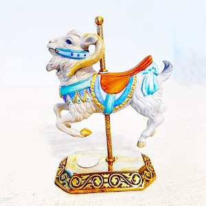 May include: A ceramic carousel ram figurine with a white body, gold horns, and blue and orange accents. The ram has a saddle and is mounted on a gold-colored pole and base. The base has an ornate design.