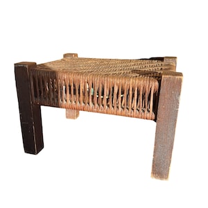 May include: A small, handcrafted wooden stool with a woven seat. The legs and frame are a dark brown, contrasting with the lighter, natural-toned woven top. The stool is designed with a simple, rustic aesthetic.