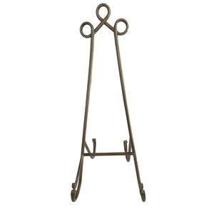 May include: A bronze-colored metal display easel. The easel has a three-legged design with decorative scrollwork at the base and top. The top features a looped design. The easel is designed to hold a picture frame or artwork.