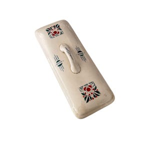 May include: Cream-colored rectangular ceramic lid with a curved handle. The lid features decorative floral and geometric patterns in red, green, and gray. The handle is a matching cream color.