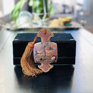 May include: An ornate, decorative pendant with a tassel. The pendant features a detailed, colorful design with red, gold, and blue accents. It is attached to a brown tassel and cord. The pendant is resting on a black box.
