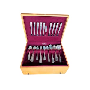 May include: A wooden silverware chest with a burgundy velvet interior. The chest is open, revealing a complete set of silver utensils, including forks, spoons, knives, and serving pieces. The silverware is neatly arranged in designated slots.