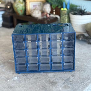 May include: A dark blue plastic storage cabinet featuring twenty-five individual clear pull-out drawers arranged in a five-by-five grid. The unit has a rectangular frame with a smooth finish and small circular openings along the top edge.