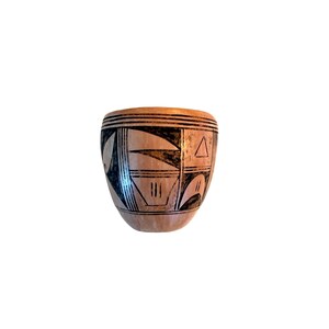 May include: A light brown ceramic pot with black geometric designs. The pot features a series of black lines and shapes, including triangles and curved lines. The pot has a rounded shape and a smooth surface.