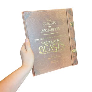 May include: A brown hardcover book titled The Case of Beasts Explore the Film Wizardry of Fantastic Beasts. The cover features gold lettering, a textured leather-like appearance, and a vertical spine-like strip with metallic brass-colored hardware.