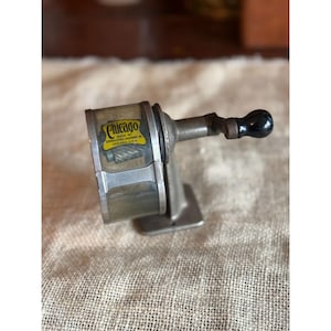 May include: Vintage Chicago pencil sharpener with a clear plastic casing and a metal body. The label reads "Chicago" and "Made by Spengler-Loomis Co. Chicago U.S.A." The sharpener has a black handle and is designed to sharpen pencils.