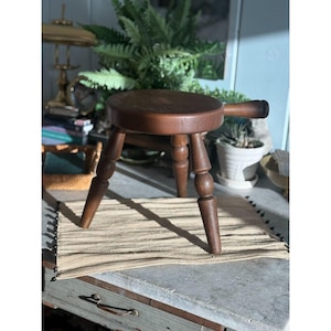 May include: A small, round, dark brown wooden stool featuring three turned legs and a single handle extending from the side of the seat. The stool is constructed from solid wood with a smooth, polished finish and a simple, functional design.