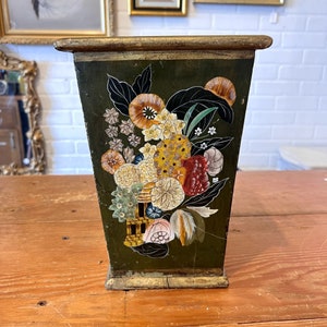 May include: A dark green, rectangular wastebasket with a floral design. The flowers are in shades of yellow, orange, red, and white, with black leaves. The top and base have a gold trim.