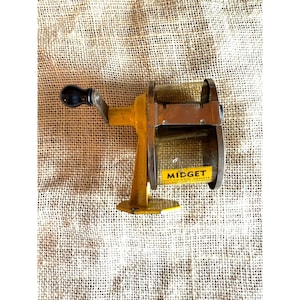 May include: A vintage yellow Midget automatic pencil sharpener with a black handle. The sharpener has a crank and a small label that reads "Midget Automatic Pencil". The sharpener is made of metal and has a rectangular shape.