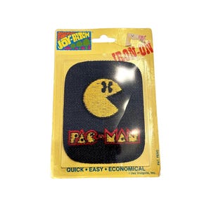 May include: A rectangular dark blue iron-on patch featuring a yellow embroidered Pac-Man character and the red and yellow text Pac-Man. The patch is sealed in yellow cardboard packaging labeled Joy-Patch, Iron-On, and Quick Easy Economical.